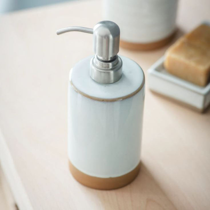 A soap dispenser with a silver metal pump, attached to a part glazed ceramic pot in Off-white with a sandy ceramic base. 