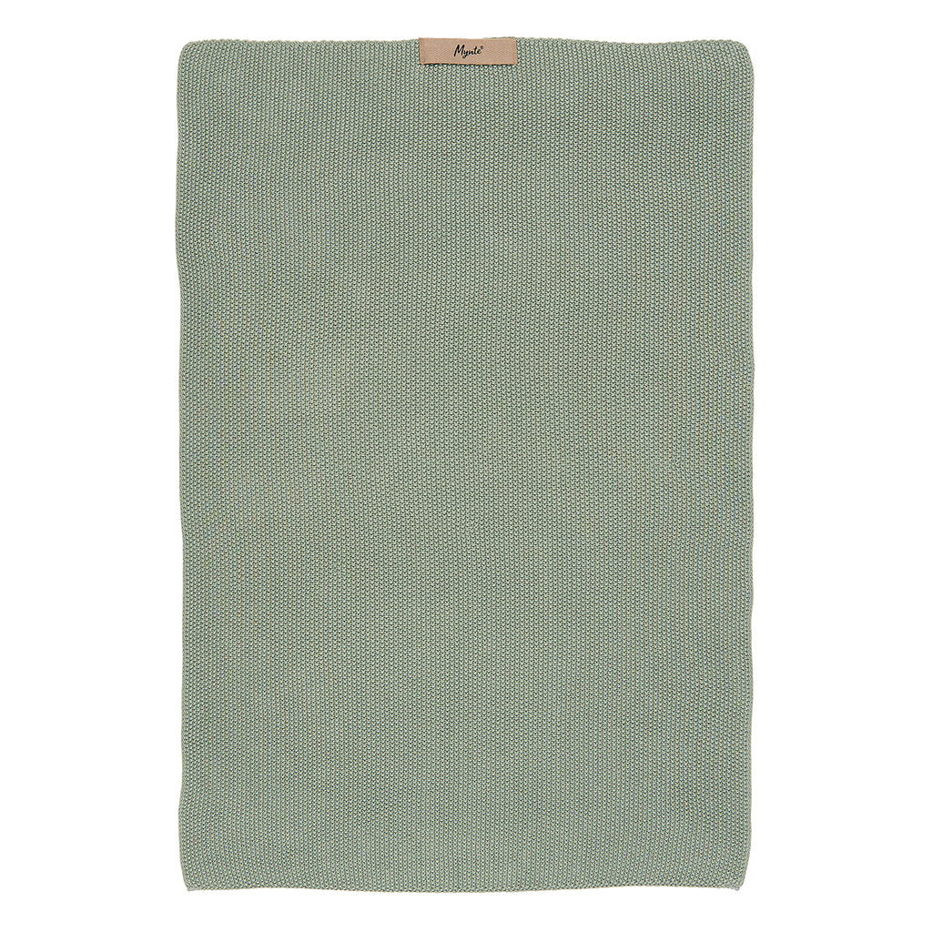 A rectangular knitted hand towel in a dusty green colour. There is a small beige coloured label at the top of the towel which says 'Mynte' in black lettering.